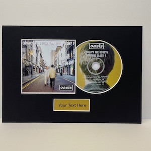 May include: A black framed CD display featuring Oasis's album "What's the Story (Morning Glory)?" The CD is displayed alongside a photo of the album cover, which shows three men walking down a city street. A gold plaque with the text "Your Text Here" is positioned below the CD.