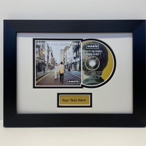 May include: A framed CD and album cover of Oasis's "What's the Story" Morning Glory? album. The album cover features a black and white photo of three men walking down a street. The CD is in a black frame with a gold plaque that says "Your Text Here".