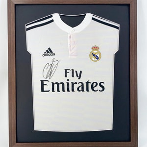 May include: A white soccer jersey with black trim and the Adidas logo. The jersey has the Real Madrid crest and the text "Fly Emirates" printed on the front. The jersey is framed in a brown wood frame.