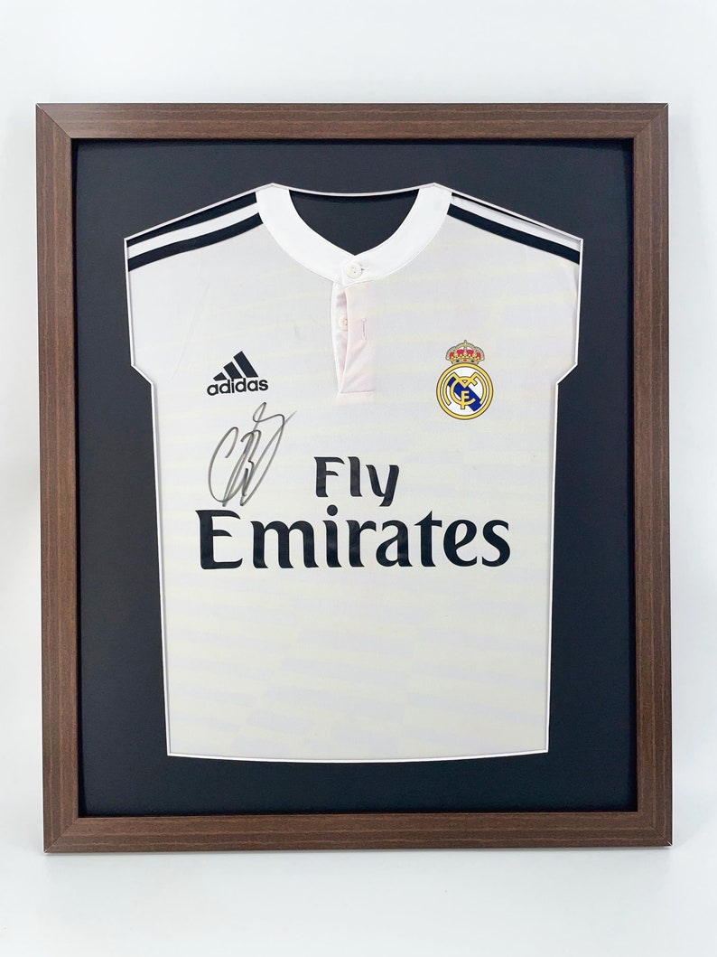 May include: A white soccer jersey with black trim and the Adidas logo. The jersey has the Real Madrid crest and the text "Fly Emirates" printed on the front. The jersey is framed in a brown wood frame.