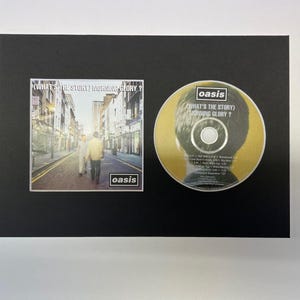 May include: A framed CD and album artwork for Oasis's "(What's the Story) Morning Glory?". The album cover depicts a street scene with two figures. The CD is gold, featuring the album title and band name.