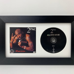 May include: A black framed picture with a white mount. The picture contains a CD and a photo of 2Pac. The CD is black with white text that reads "2Pac All Eyez On Me Disc 1". The photo is of 2Pac with the text "2Pac All Eyez On Me" above him.