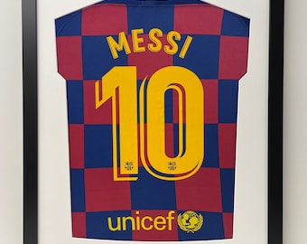 May include: A framed soccer jersey with a red and blue checkered pattern. The jersey has the name "MESSI" and the number "10" on the back. The jersey also has the UNICEF logo and the text "unicef" on the bottom.