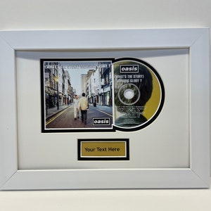 May include: A white framed picture featuring an Oasis CD and album cover for "What's the Story" Morning Glory?. The album cover shows a street scene with three people walking down the street. The CD is in a black frame with a white border. The text "Your Text Here" is on a gold plaque.
