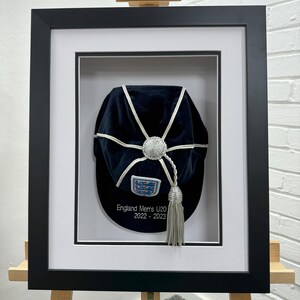 May include: A framed black cap with white detailing and a silver tassel. The cap features a crest and the text "England Men's U20 2022-2023". The frame is black with a white mount, displayed on a wooden easel.