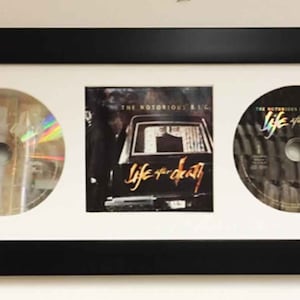 May include: A black framed triple matted display featuring three items: two silver CDs with the text "Life After Death" and a CD cover with the text "The Notorious B.I.G. Life After Death"