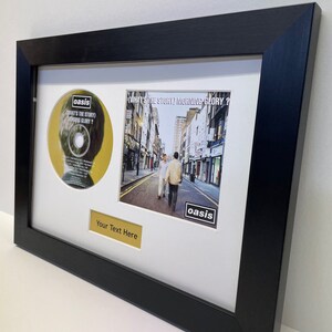 May include: A black framed picture with a white mount. The picture includes a CD and a photo of a street scene. The CD is yellow with the text "What's the Story (Morning Glory?)" and the word "oasis" in a black box. The photo shows two people walking down a street with buildings on either side. The photo also has the word "oasis" in a black box. The text "Your Text Here" is on a gold plaque below the photo.