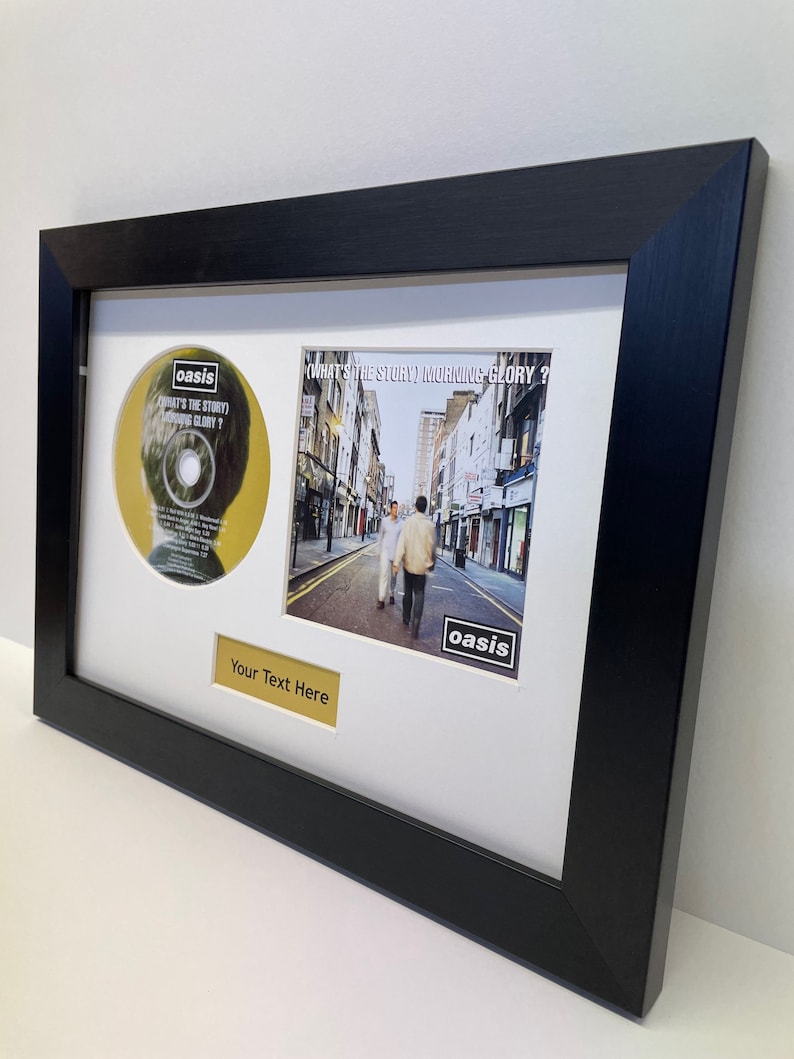 May include: A black framed picture with a white mount. The picture includes a CD and a photo of a street scene. The CD is yellow with the text "What's the Story (Morning Glory?)" and the word "oasis" in a black box. The photo shows two people walking down a street with buildings on either side. The photo also has the word "oasis" in a black box. The text "Your Text Here" is on a gold plaque below the photo.