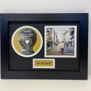 May include: A black framed picture featuring a CD and album cover by the band Oasis. The CD is titled "What's the Story (Morning Glory?)" and the album cover depicts three people walking down a street. The text "Your Text Here" is on a gold plaque beneath the album cover.