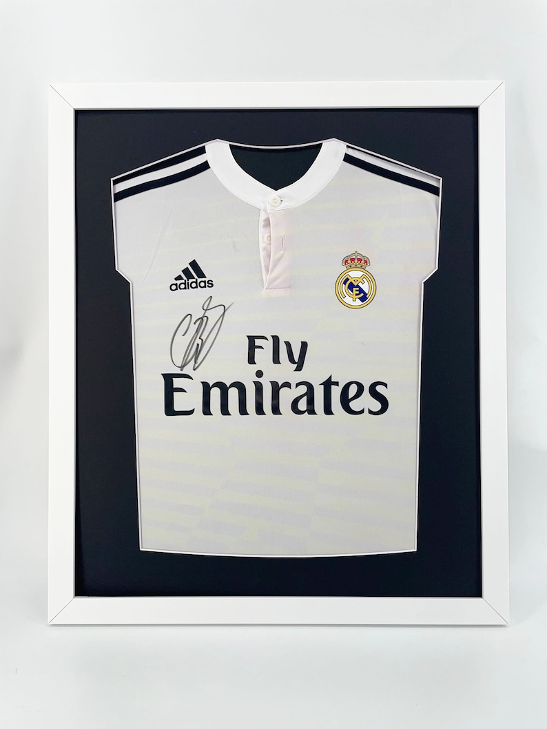 May include: A white soccer jersey with black stripes on the shoulders and a black Adidas logo. The jersey has the Real Madrid crest and the text "Fly Emirates" on the front. The jersey is signed with a black signature.