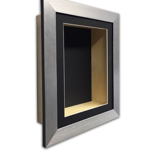 May include: A shadow box with a brushed silver frame and a black interior. The box has a rectangular opening with a black border and a light wood-coloured interior. The shadow box is designed to display small objects.