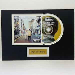 May include: A black framed CD display with a white mount and a gold plaque. The CD is a yellow and white disc with the text "(What's the Story) Morning Glory?" and "oasis" printed on it. The plaque has the text "Your Text Here" printed on it.