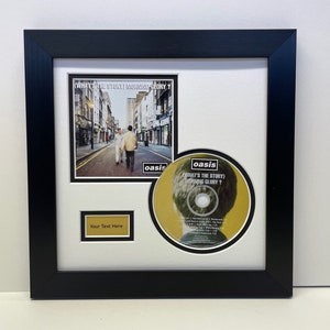 May include: A framed CD and album cover set featuring Oasis's "What's the Story (Morning Glory)?" album. The album cover features a black and white photo of a street scene with three people walking down the street. The CD is yellow with a black and white photo of a person's head on it. The frame is black with a white mount.