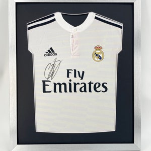 May include: A white soccer jersey with black trim and the Adidas logo. The jersey has the Real Madrid crest and the text "Fly Emirates" on the front. The jersey is framed in a silver frame with a black mat.