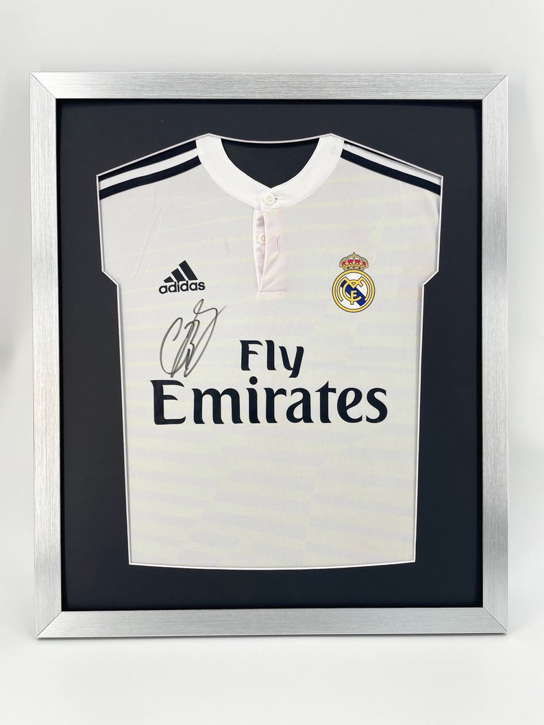 May include: A white soccer jersey with black trim and the Adidas logo. The jersey has the Real Madrid crest and the text "Fly Emirates" on the front. The jersey is framed in a silver frame with a black mat.