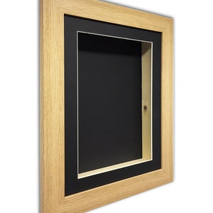 May include: A rectangular shadow box frame with a natural wood finish. The frame features a black interior and a white border, creating a layered effect. The shadow box is designed for displaying objects and artwork.
