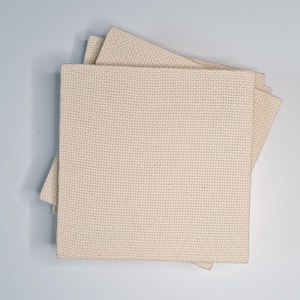 Op de afbeelding: Een stapel van vier vierkante, gebroken witte kruissteekstofstukken. De stof heeft een rasterpatroon van kleine gaatjes, perfect voor handwerken. De stukken zijn gelaagd, waarbij het bovenste stuk iets verschoven is, waardoor de randen zichtbaar zijn.