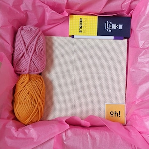 Könnte beinhalten: Ein Bastelset in einer rosa Box mit rosa Seidenpapier. Das Set enthält rosa und orangefarbenes Garn, eine Leinwand, einen Stift und eine gelbe Box mit dem Text "PUNCH NEEDLE". Ein kleines gelbes Quadrat mit dem Text "oh!" ist ebenfalls enthalten.