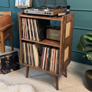 May include: A wooden record player stand with two shelves for vinyl storage, featuring cane side panels and four tapered legs. The top surface holds a turntable with a clear dust cover, while the shelves are filled with numerous vinyl records.