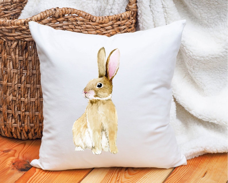 May include: White cushion with a watercolour illustration of a brown rabbit with pink ears. The rabbit is sitting on its hind legs and looking to the left.