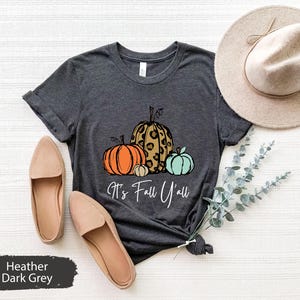 Peut inclure: Un t-shirt gris foncé chiné avec un motif de trois citrouilles. Les citrouilles sont orange, marron avec un imprimé léopard et turquoise. Le texte "It's Fall Y'all" est imprimé sous les citrouilles.