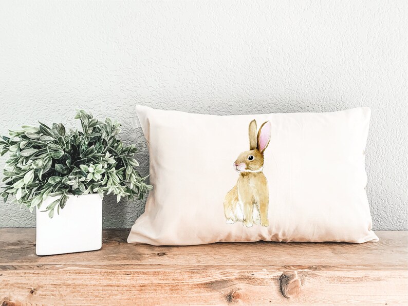 May include: A beige pillow with a watercolour illustration of a brown rabbit with pink ears.