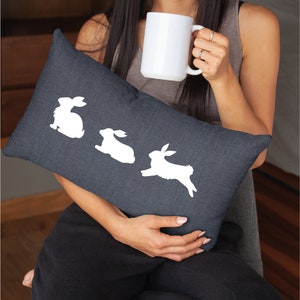 May include: A dark gray throw pillow with white bunny silhouettes. The pillow is being held by a person.