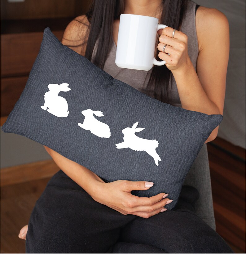 May include: A dark gray throw pillow with white bunny silhouettes. The pillow is being held by a person.