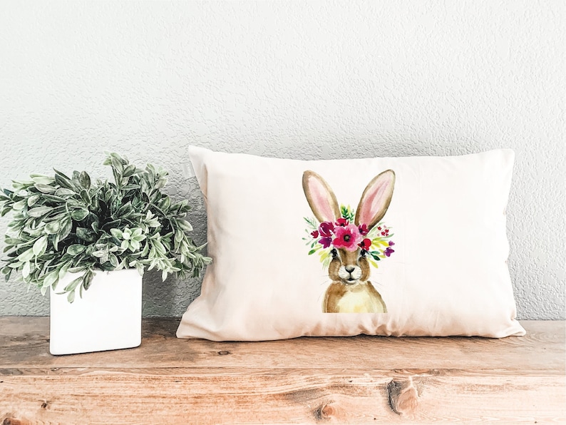 May include: A white pillow with a brown bunny wearing a pink and purple flower crown. The bunny is looking at the camera.