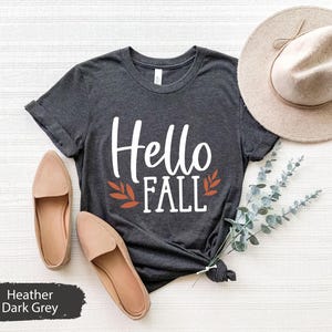 May include: A dark heather grey t-shirt with the text "Hello Fall" in white with orange leaf accents.