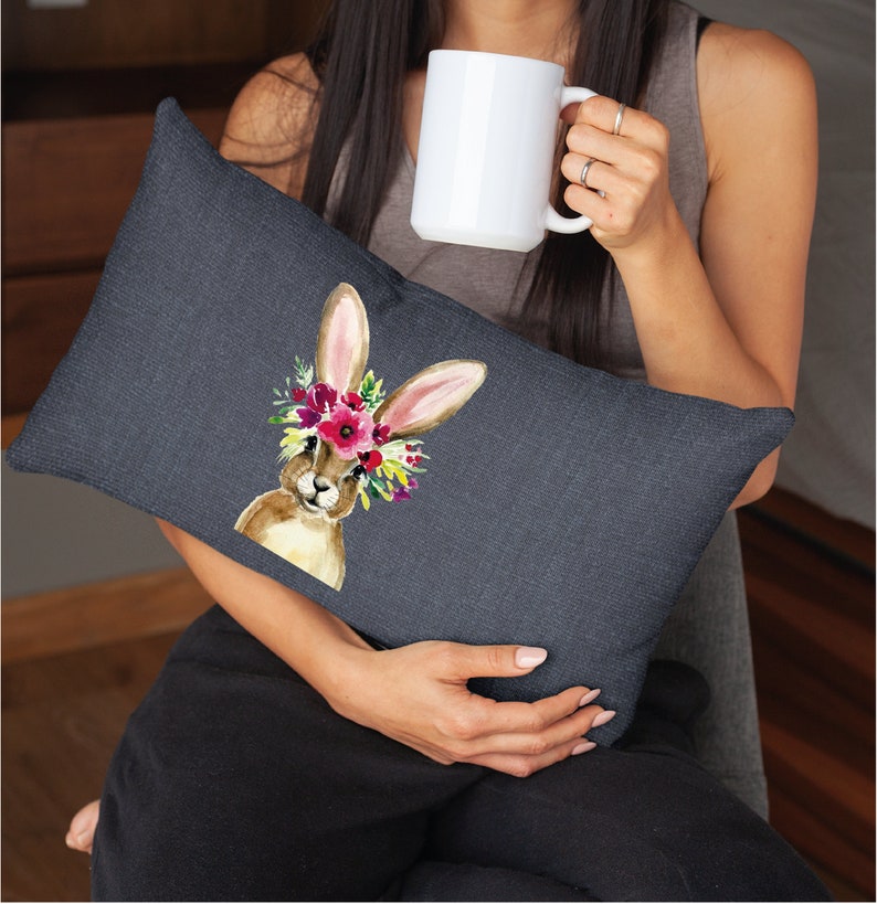 May include: A dark gray throw pillow with a watercolor illustration of a brown bunny wearing a floral crown. The bunny is looking directly at the viewer.