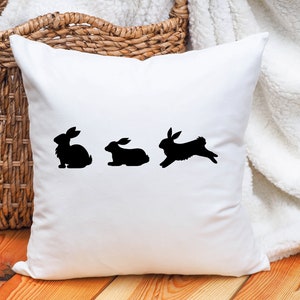 May include: White throw pillow with three black bunny silhouettes. The bunnies are in a line, with the first bunny sitting, the second bunny sitting, and the third bunny hopping.