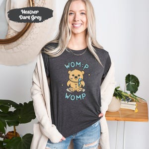 Può includere: Una t-shirt color grigio scuro melange con un orso dei cartoni animati che piange e il testo "WOMP" sopra e sotto l'orso.