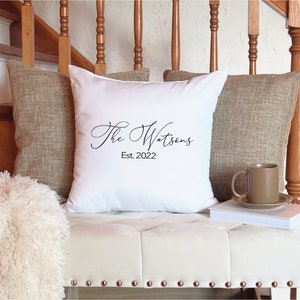 May include: A white decorative pillow with the text "The Watsons Est. 2022" in black script. The pillow is flanked by two brown textured pillows and rests on a white bench with metal studs. A brown mug and saucer sit on a stack of books.