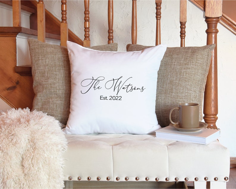 May include: A white decorative pillow with the text "The Watsons Est. 2022" in black script. The pillow is flanked by two brown textured pillows and rests on a white bench with metal studs. A brown mug and saucer sit on a stack of books.