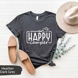 Könnte beinhalten: Dunkelgraues T-Shirt mit dem Text "Happy Camper" in einem weißen Rechteck mit einer Grafik einer Strichmännchenfigur, die ein Marshmallow über einem Lagerfeuer hält.