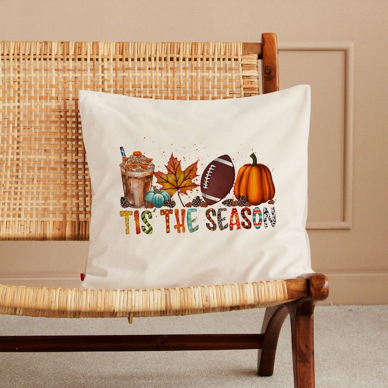 May include: A white throw pillow with a colorful graphic design featuring a football, pumpkin, fall leaves, and a cup of coffee. The text "Tis the Season" is written in colorful letters.