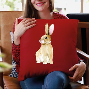 May include: A red cushion with a watercolour illustration of a brown rabbit with pink ears. The rabbit is sitting on its hind legs and looking to the left.