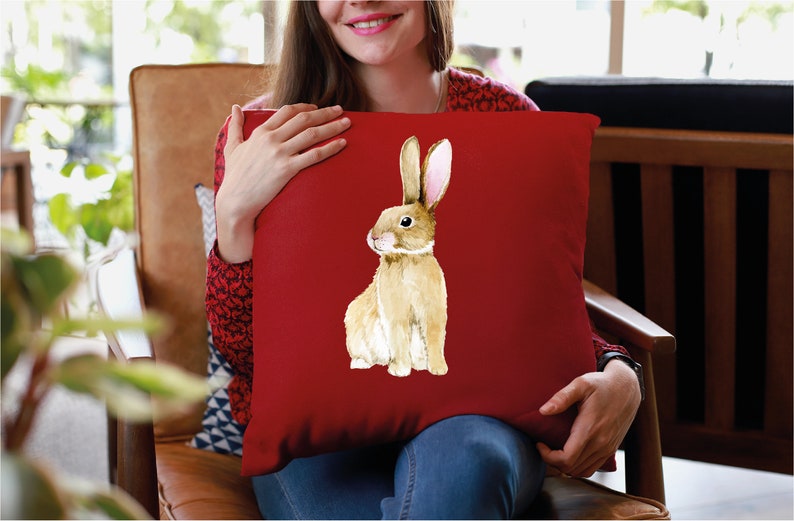 May include: A red cushion with a watercolour illustration of a brown rabbit with pink ears. The rabbit is sitting on its hind legs and looking to the left.