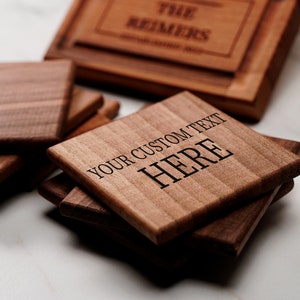 May include: A stack of four square wooden coasters with a dark brown wood grain. The top coaster has the text "YOUR CUSTOM TEXT HERE" engraved on it.