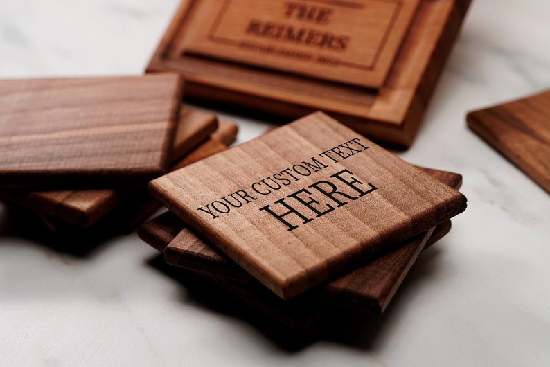 May include: A stack of four square wooden coasters with a dark brown wood grain. The top coaster has the text "YOUR CUSTOM TEXT HERE" engraved on it.