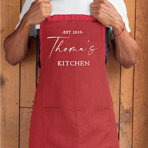 May include: A red kitchen apron with a white pocket and the text "EST 2010 - Thomas' Kitchen" printed on the front.