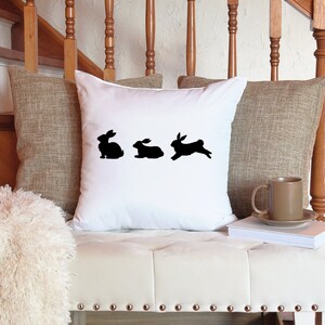 May include: White throw pillow with a black silhouette of three rabbits, one standing and two hopping.