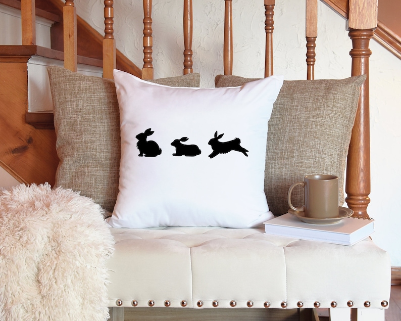 May include: White throw pillow with a black silhouette of three rabbits, one standing and two hopping.