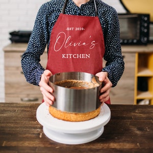 May include: A person wearing a red apron with the text "EST 2010 - Olivia's Kitchen" is holding a round cake pan over a cake on a white cake stand.