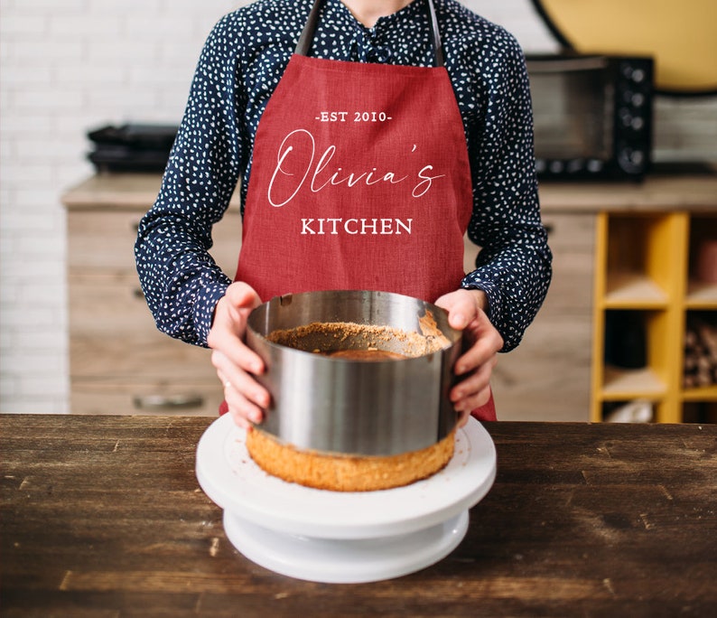 May include: A person wearing a red apron with the text "EST 2010 - Olivia's Kitchen" is holding a round cake pan over a cake on a white cake stand.