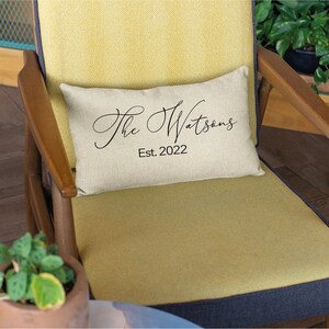 May include: A rectangular beige pillow with the text "The Watsons" in elegant black script and "Est. 2022" below. The pillow rests on a yellow chair with wooden arms and a dark blue seat.