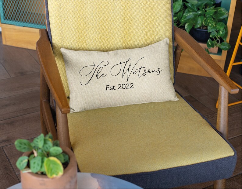 May include: A rectangular beige pillow with the text "The Watsons" in elegant black script and "Est. 2022" below. The pillow rests on a yellow chair with wooden arms and a dark blue seat.