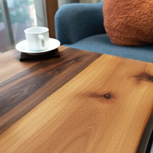 Op de afbeelding: Een close-up van een houten tafelblad met een lichtbruin en donkerbruin houtnerfpatroon. Het tafelblad heeft een natuurlijke rand en een klein knoopje in het hout. Een witte koffiekop en een schoteltje staan op de tafel.