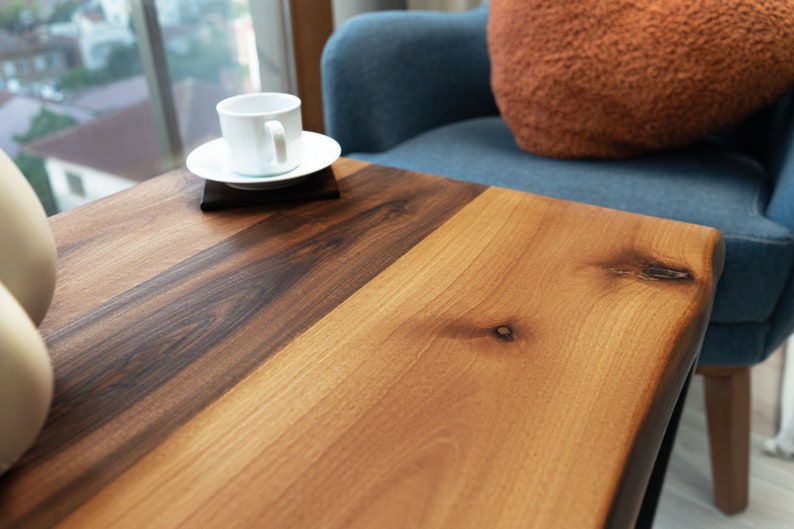 Op de afbeelding: Een close-up van een houten tafelblad met een lichtbruin en donkerbruin houtnerfpatroon. Het tafelblad heeft een natuurlijke rand en een klein knoopje in het hout. Een witte koffiekop en een schoteltje staan op de tafel.